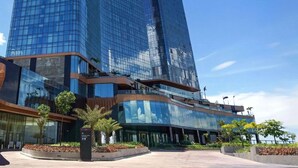 A modern hotel with a glass facade, a lower level with a curved roof, and a landscaped area with palm trees.