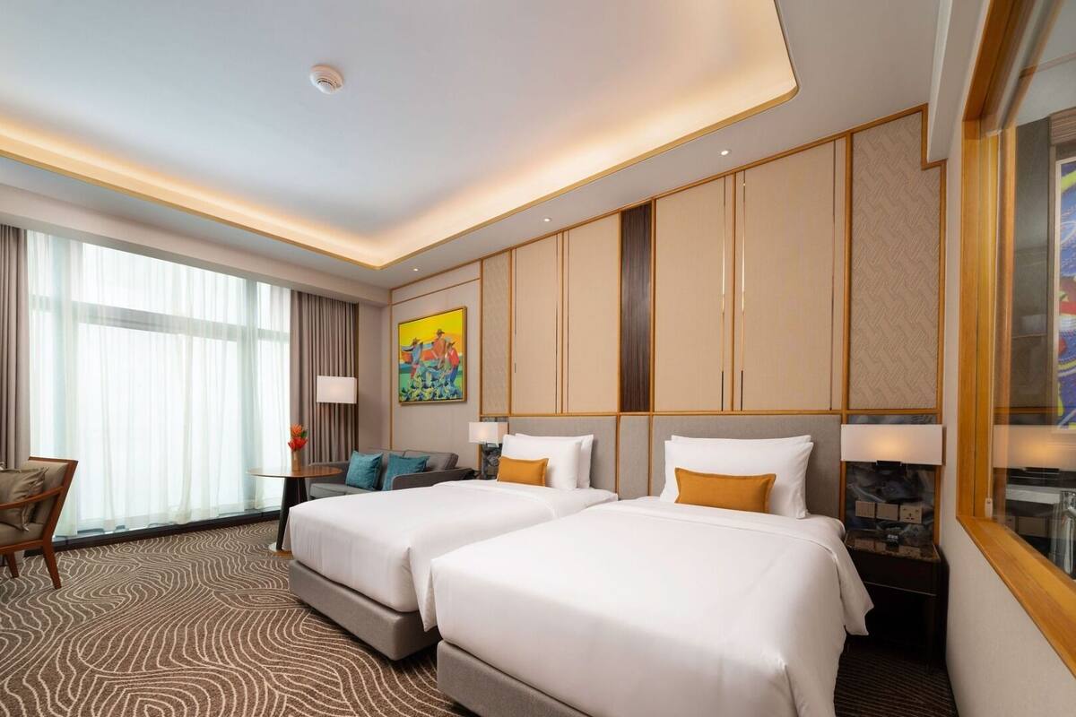 A hotel room with two beds, a desk, a chair, a large window, and a painting on the wall.