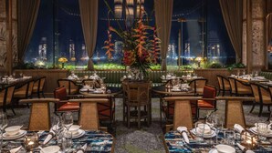 Un restaurante con varias mesas dispuestas para comensales, amplias ventanas que ofrecen vistas de la ciudad y un candelabro moderno colgado del techo.