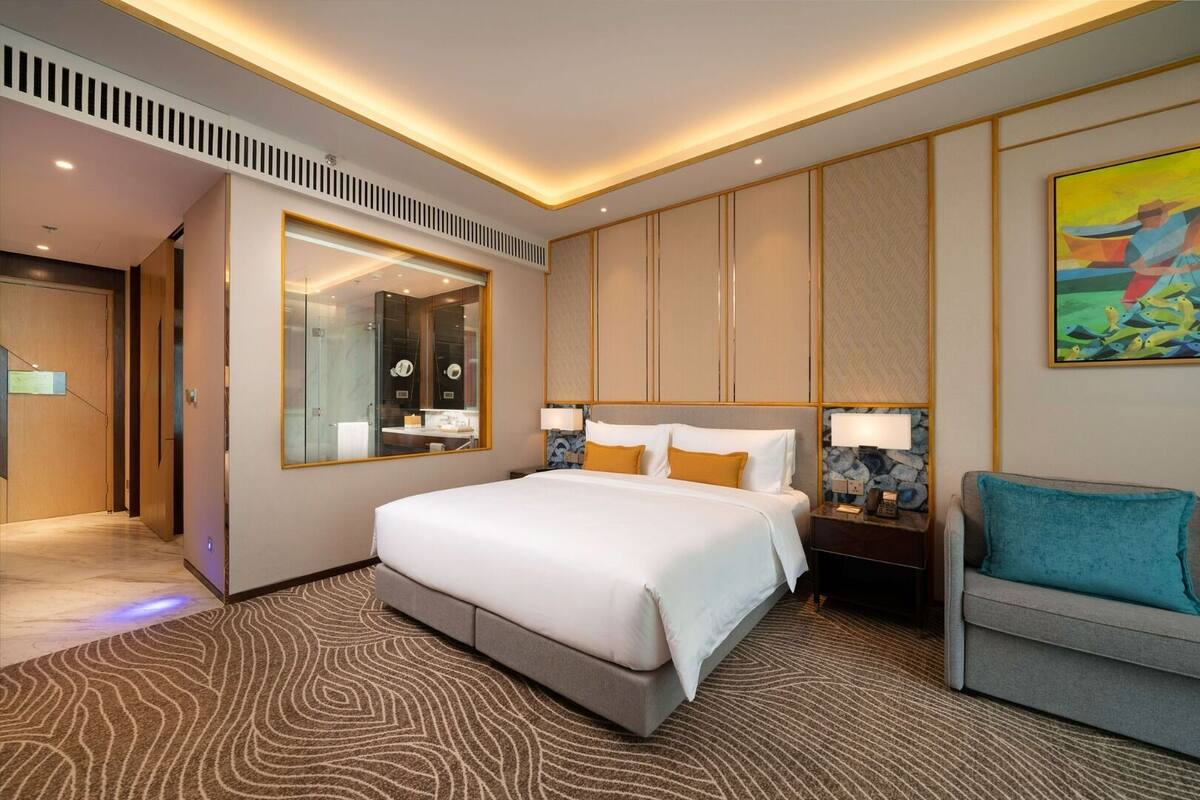 A modern hotel room with a large bed, a seating area, a mirror, and a painting on the wall.