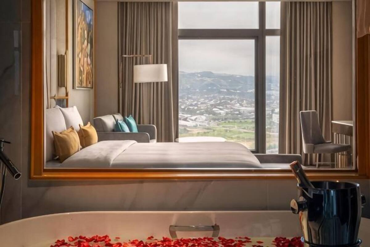A hotel room with a large bed, a view of a cityscape, a bathtub with rose petals, and a chair by the window.