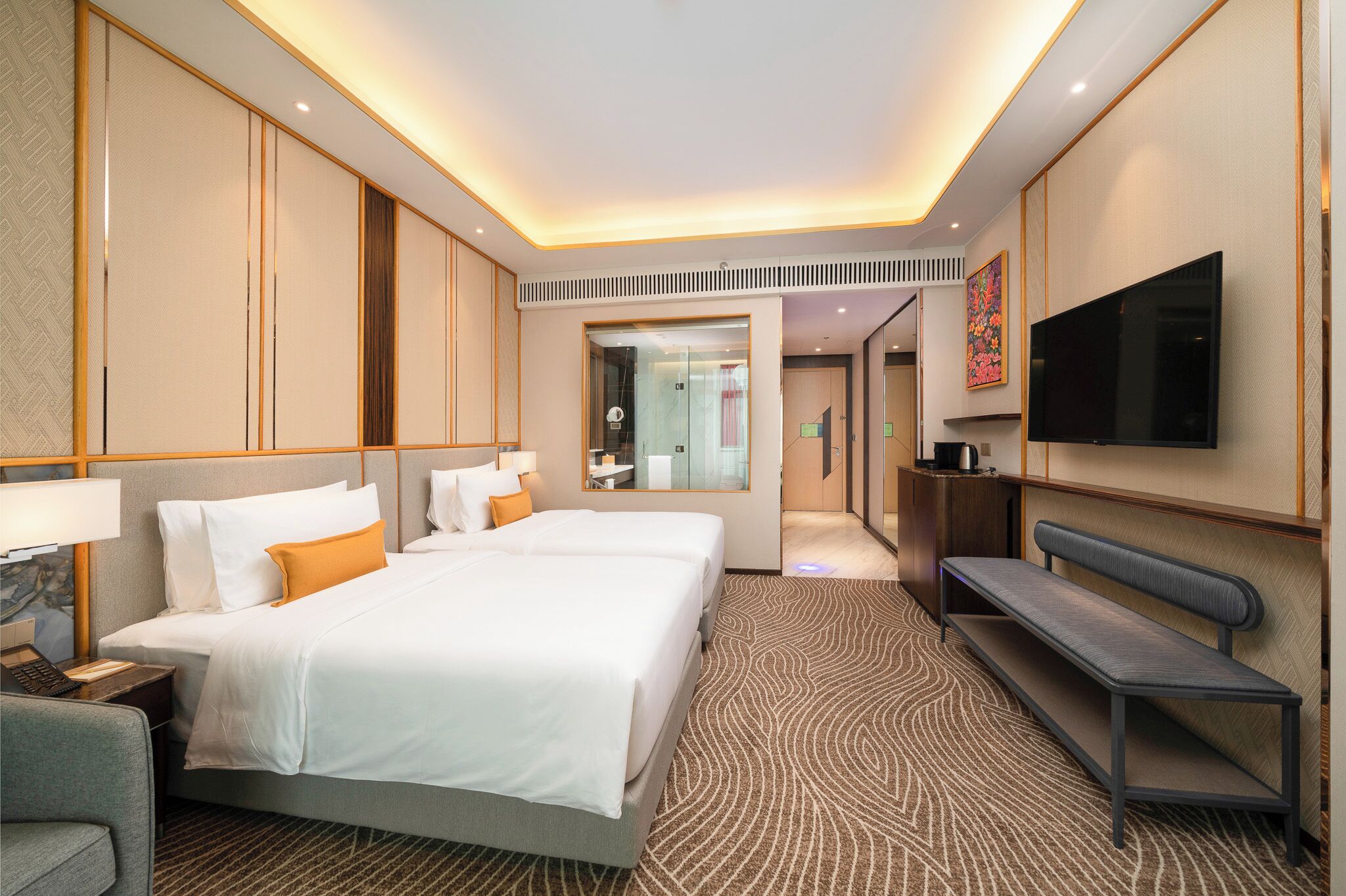 A hotel room with two beds, a bench, a TV, and a bathroom visible through a glass door.