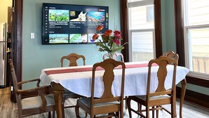 Spisestue med et bord dækket til fire personer, træstole, en vase med blomster og et fladskærms-tv på væggen, der viser et computerspil.