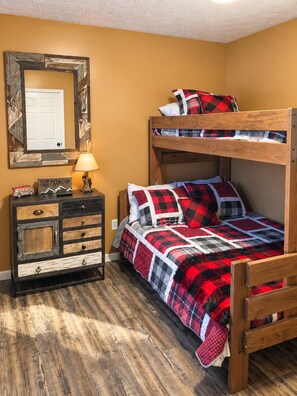 Un lit superposé avec une couverture à carreaux rouges et noirs, une commode en bois avec de nombreux tiroirs, un miroir avec un cadre rustique et une lampe posée sur la commode.