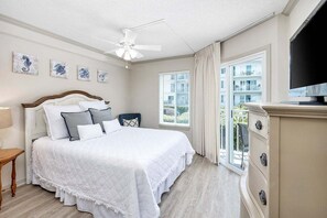 A bedroom with a large bed, a ceiling fan, a TV, and a window with a view of buildings. - Spacious and Oceanfront! (Saint Simons Island)