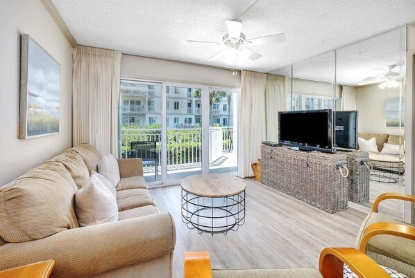 A modern living room with a sofa, a coffee table, a TV, and a balcony with a view of other buildings. - Spacious and Oceanfront! (Saint Simons Island)