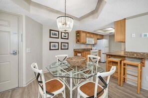 A modern kitchen with a glass dining table, white chairs, wooden cabinets, and a chandelier.
