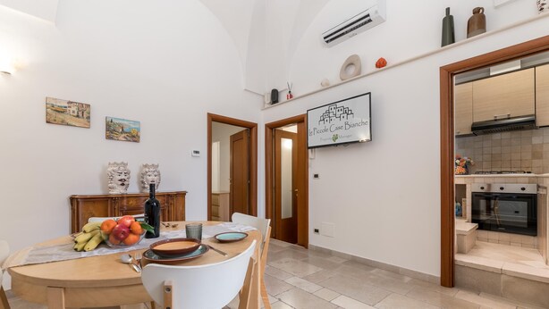 Un coin repas avec une table en bois, des chaises et un panier de fruits. Il y a deux portes qui donnent sur d’autres pièces, ainsi qu’une cuisine équipée d’une cuisinière et d’un four.