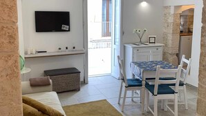 A cozy living area with a sofa, a dining table with chairs, a TV mounted on the wall, and a kitchen visible through an open door. - biancostuni66, Le Piccole Case Bianche (Ostuni)