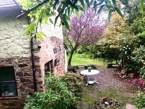 A rustic stone building with a small garden area featuring a round table and benches, surrounded by flowering plants and trees. - Garden Cottage, Wiveliscombe (Wiveliscombe)