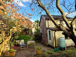A rustic stone house with a garden, wooden furniture, and a water tank. - Garden Cottage, Wiveliscombe (Wiveliscombe)