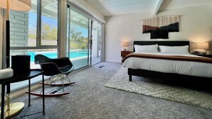 Quarto com cama, escrivaninha, cadeira, uma janela grande e vista para uma piscina.