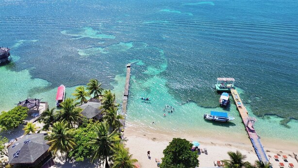 Vista aerea di un resort sulla spiaggia con acque cristalline turchesi, un molo e diverse imbarcazioni.