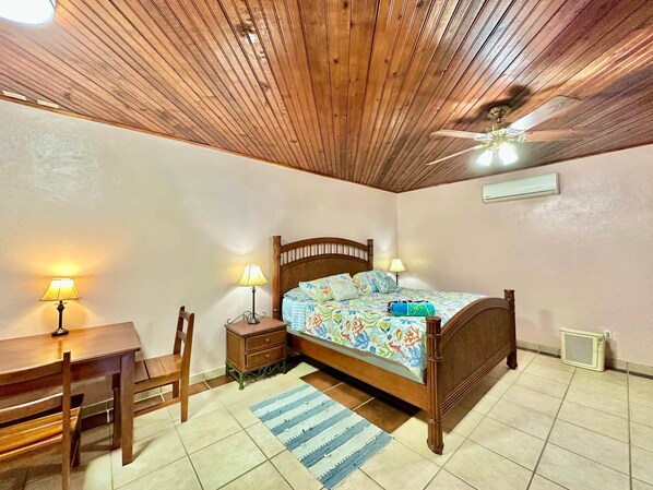 Une chambre avec un plafond en bois, un lit recouvert d’une literie à motifs floraux, deux lampes de chevet, un bureau avec une chaise et un petit tapis.