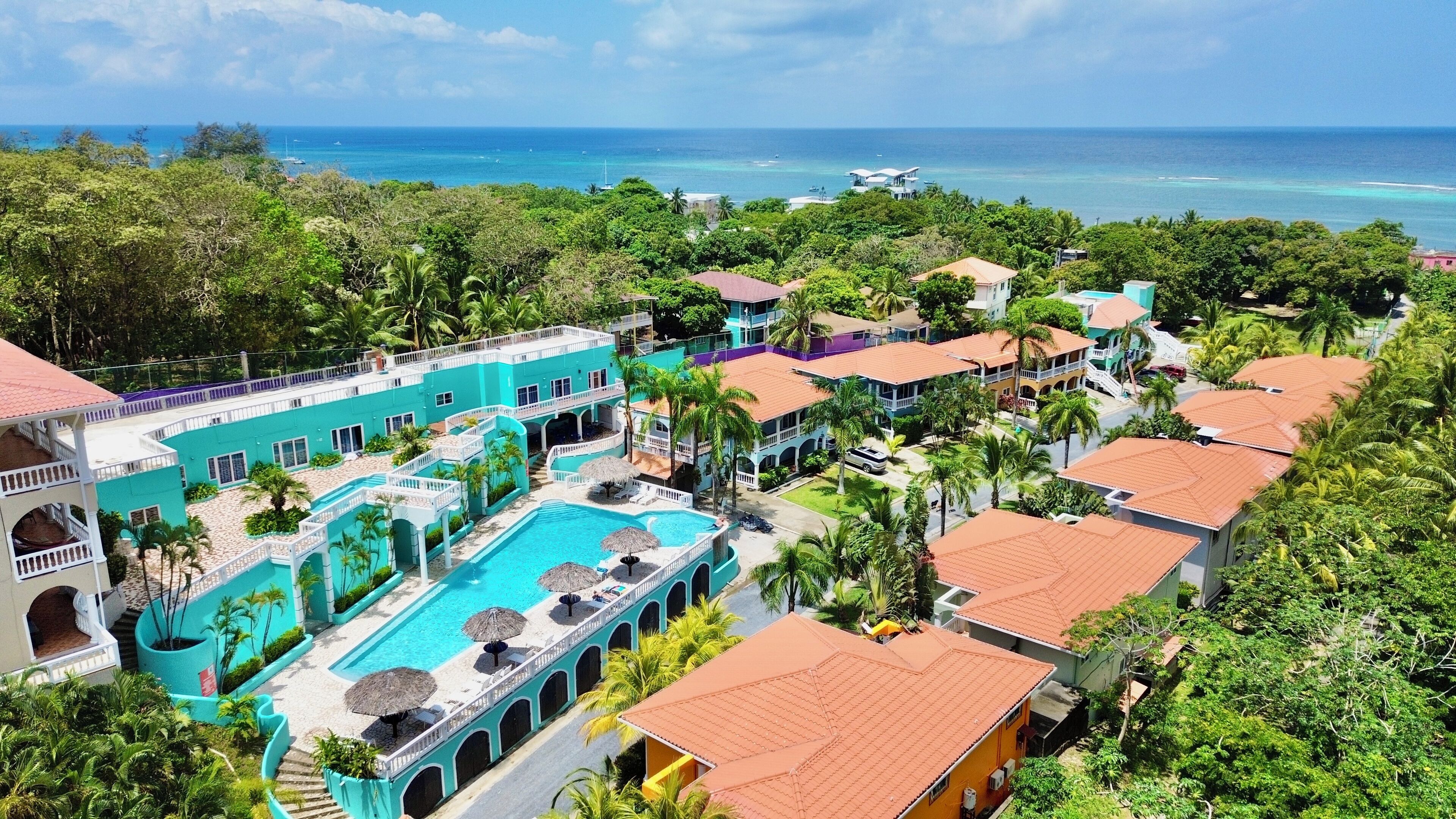 A resort with a central swimming pool, multiple buildings, and a view of the ocean.