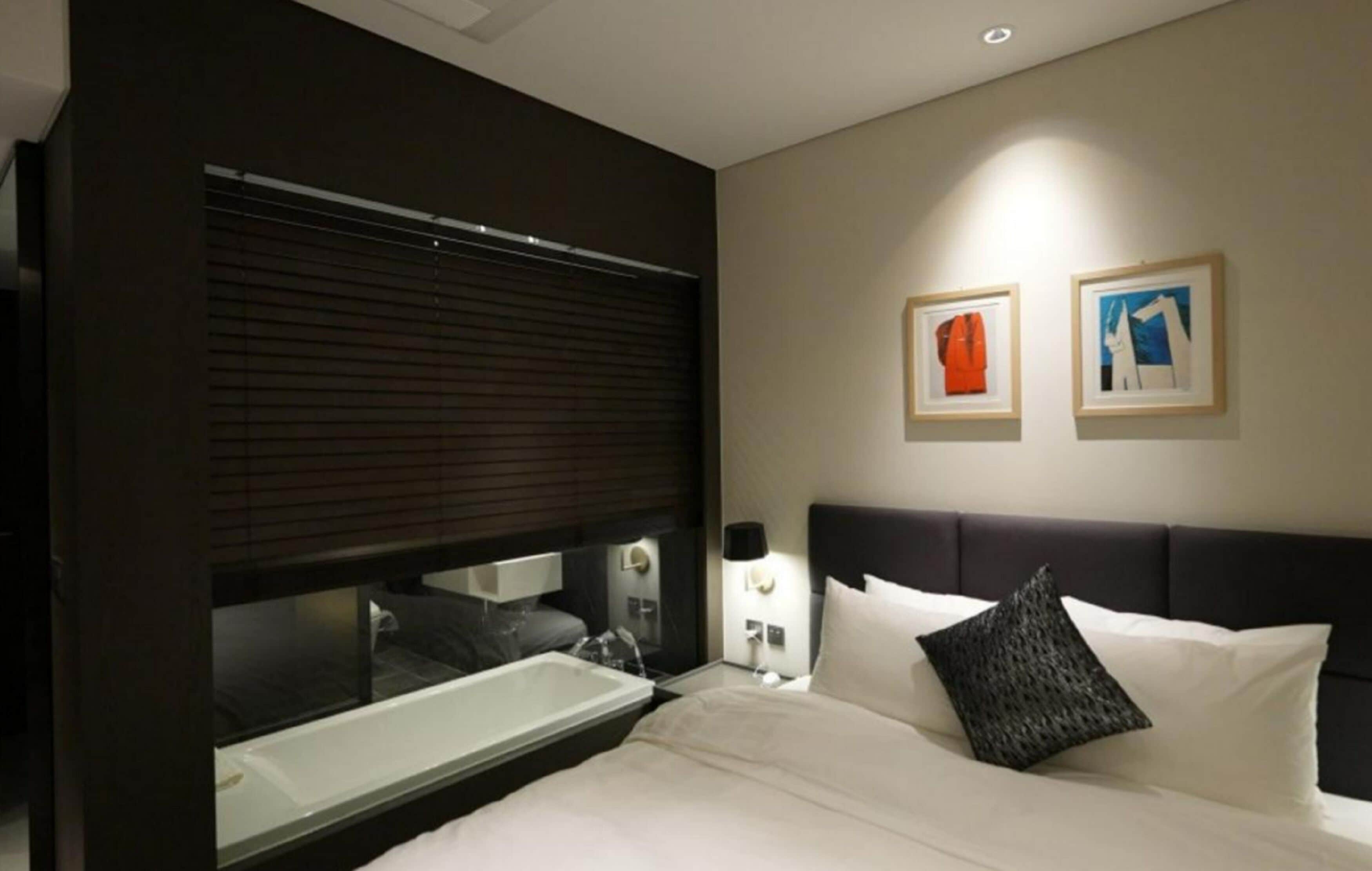 A modern hotel room with a large bed, a built-in window with blinds, a bedside table with a lamp, and two framed artworks on the wall.