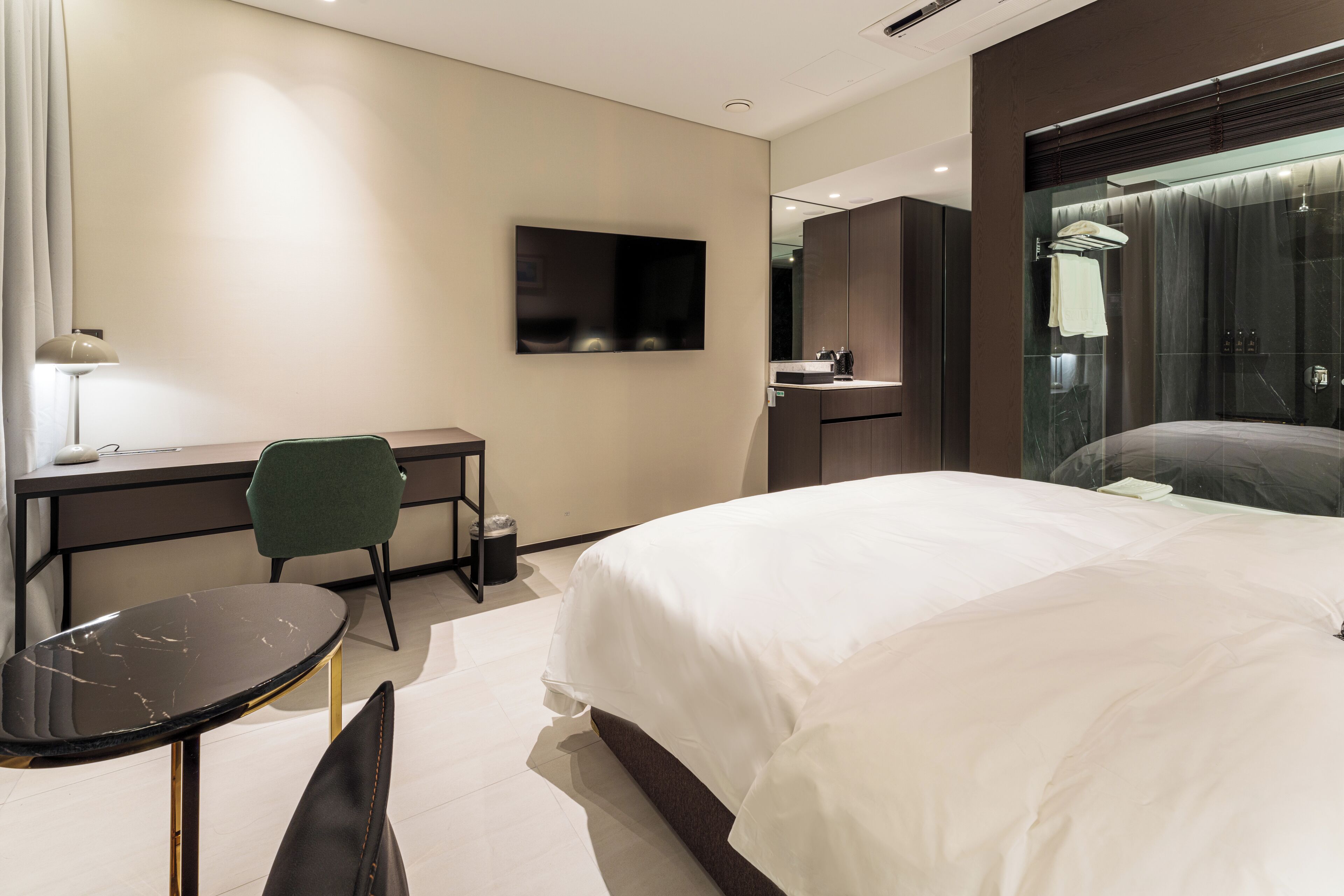 A modern hotel room with a large bed, a desk with a chair, a TV mounted on the wall, and a bathroom with a glass shower.