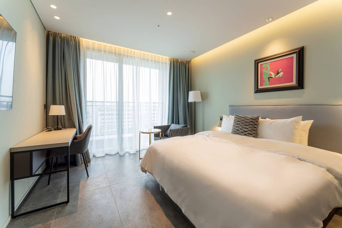 A hotel room with a large bed, a desk with a chair, a small table, and a lamp.