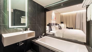 A modern bathroom with a large mirror, a white sink, a bathtub, and a large mirror reflecting a bedroom with a bed and curtains.