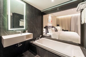 A modern bathroom with a large mirror, a white sink, a bathtub, and a large mirror reflecting a bedroom with a bed and curtains.