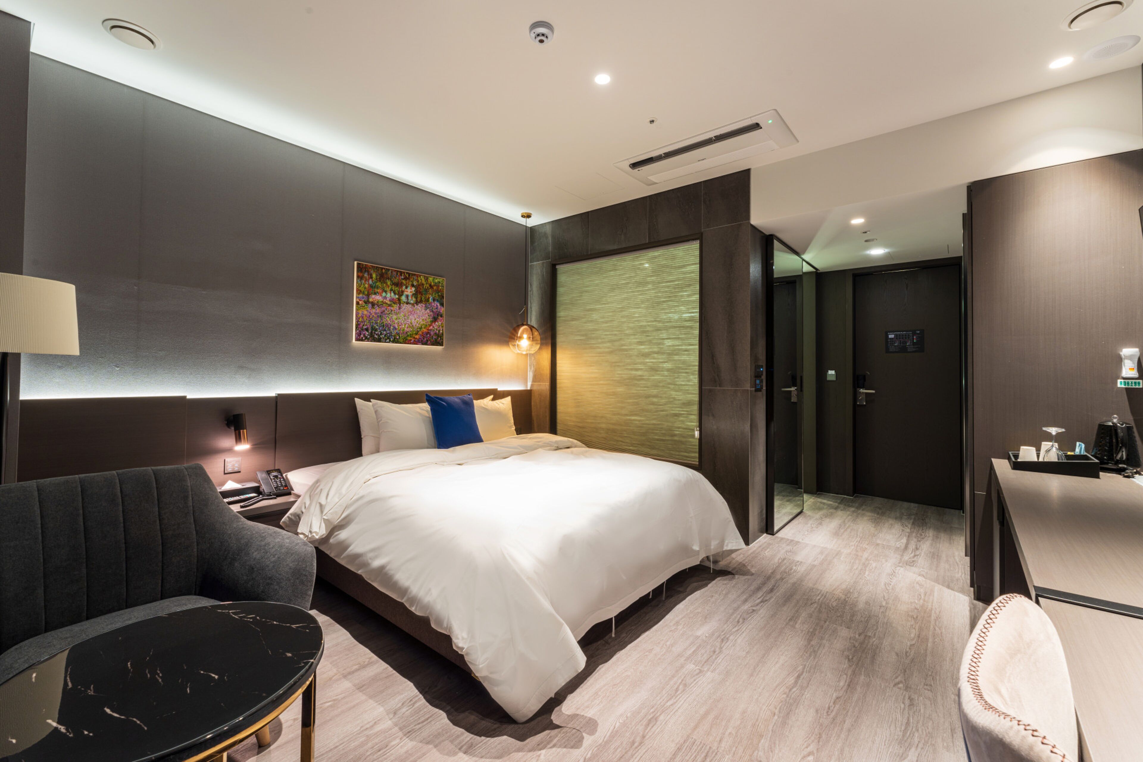 A modern hotel room with a large bed, a desk, a chair, a lamp, and a painting on the wall.