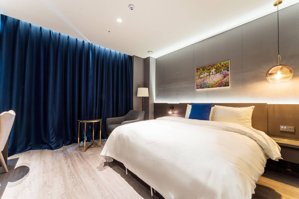 A modern hotel room with a large bed, a desk, a chair, and a painting on the wall.