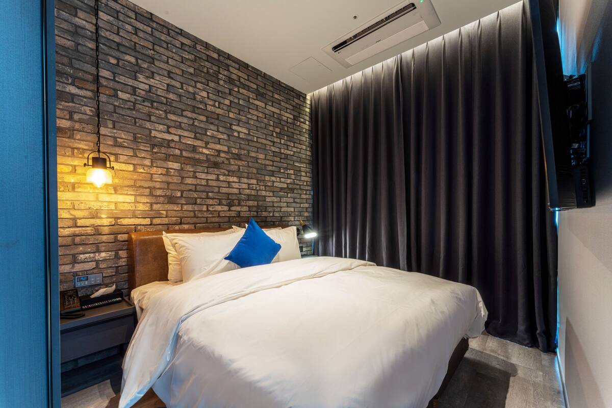 A modern hotel room with a brick wall, a large bed with white bedding, a blue pillow, a bedside lamp, and a dark curtain.
