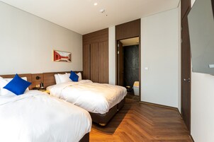 Habitación de hotel con dos camas, piso de madera, un cuadro en la pared y una puerta que da a otra habitación.