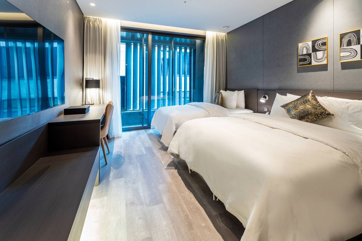 A modern hotel room with a large bed, a desk, and a view of the city through floor-to-ceiling windows.