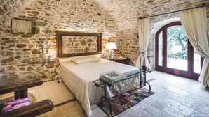 Ein Schlafzimmer mit Steinsandsteinwand, einem Bett mit Kopfteil, einem Nachttisch, einer Lampe und Blick ins Freie durch ein Fenster.