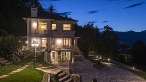 A two-story house with a stone pathway leading to the entrance, surrounded by greenery and mountains in the background. - VILLA NINFEA DEL LAGO (Baveno)