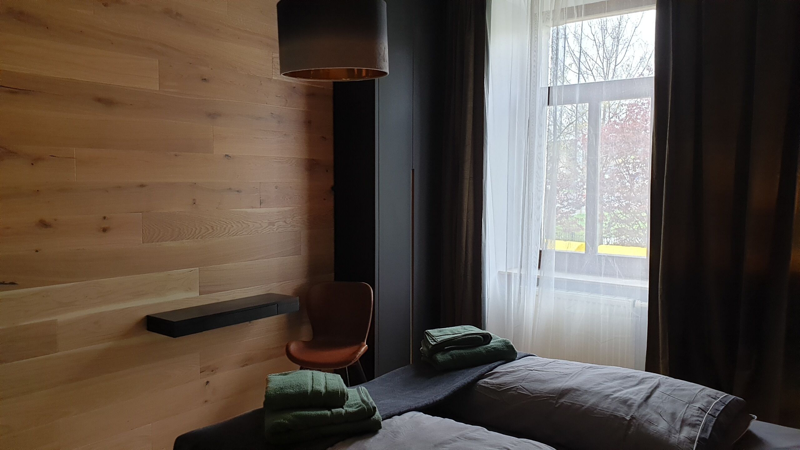 Een kamer met een houten wand, een stoel, een bed met dekbed en een raam met gordijnen.