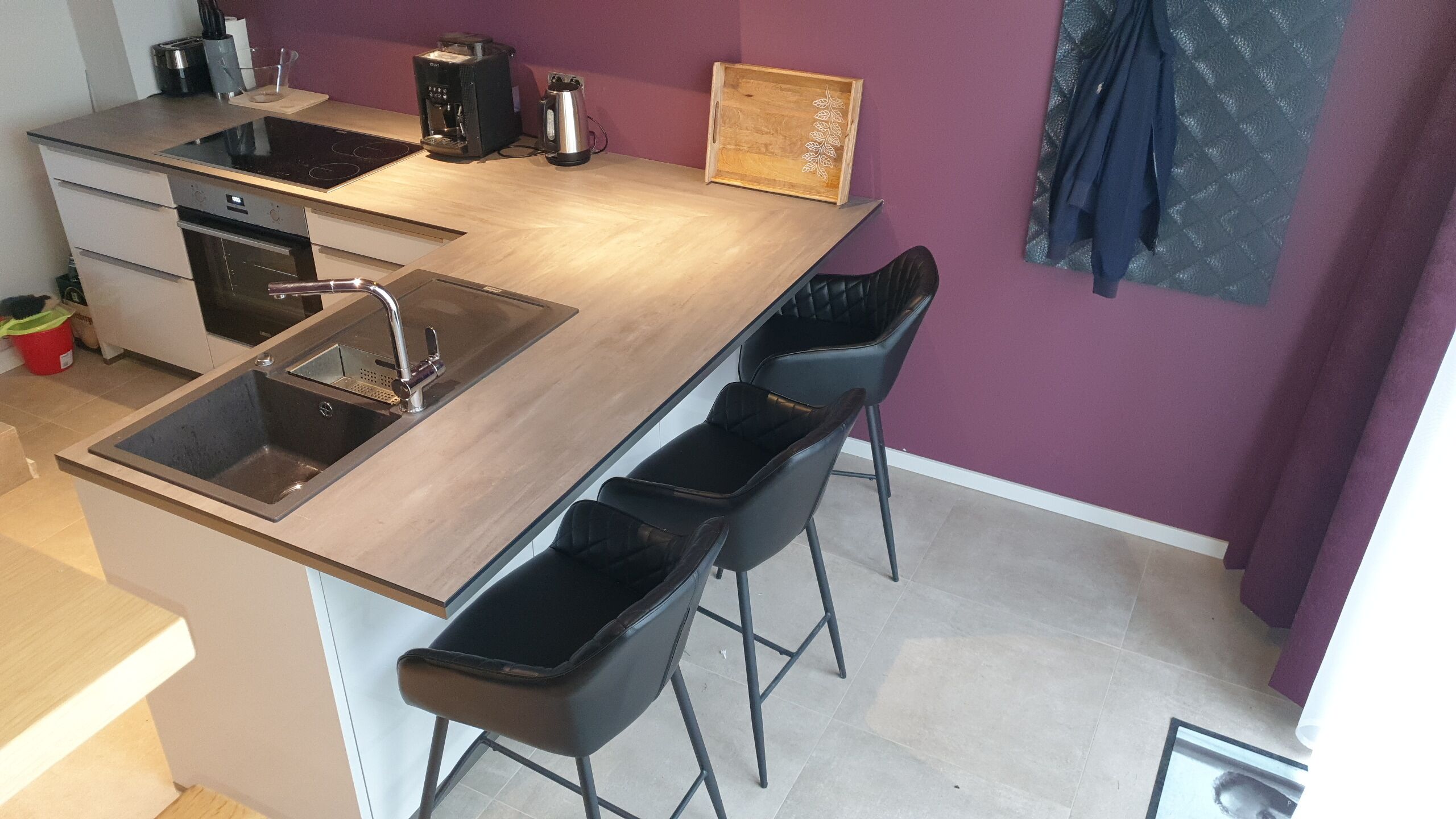 Moderni keittiö, jossa on integroitu pesuallas, kahvinkeitin ja vedenkeitin. Keittiössä on kaksi mustaa baarijakkaraa ja puinen sivupöytä.