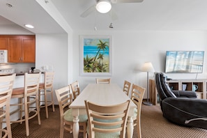 Uma cozinha moderna com armários de madeira, uma mesa de jantar com cadeiras, um sofá de couro preto, uma pintura de uma praia e uma televisão sobre um móvel.