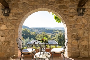 A stone archway leading to a balcony with a table and chairs, overlooking a forested landscape.