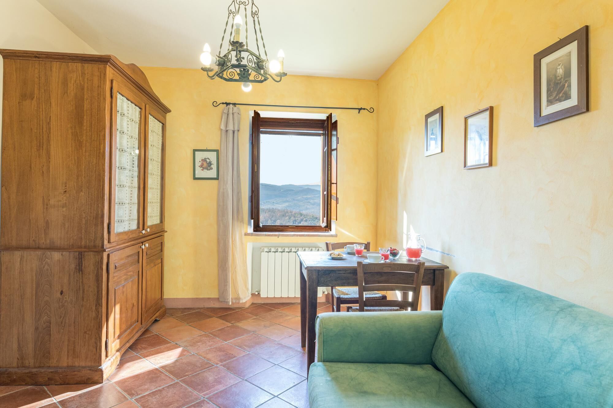 A cozy room with a wooden cabinet, a dining table with chairs, a green sofa, and a window with a view of the hills.