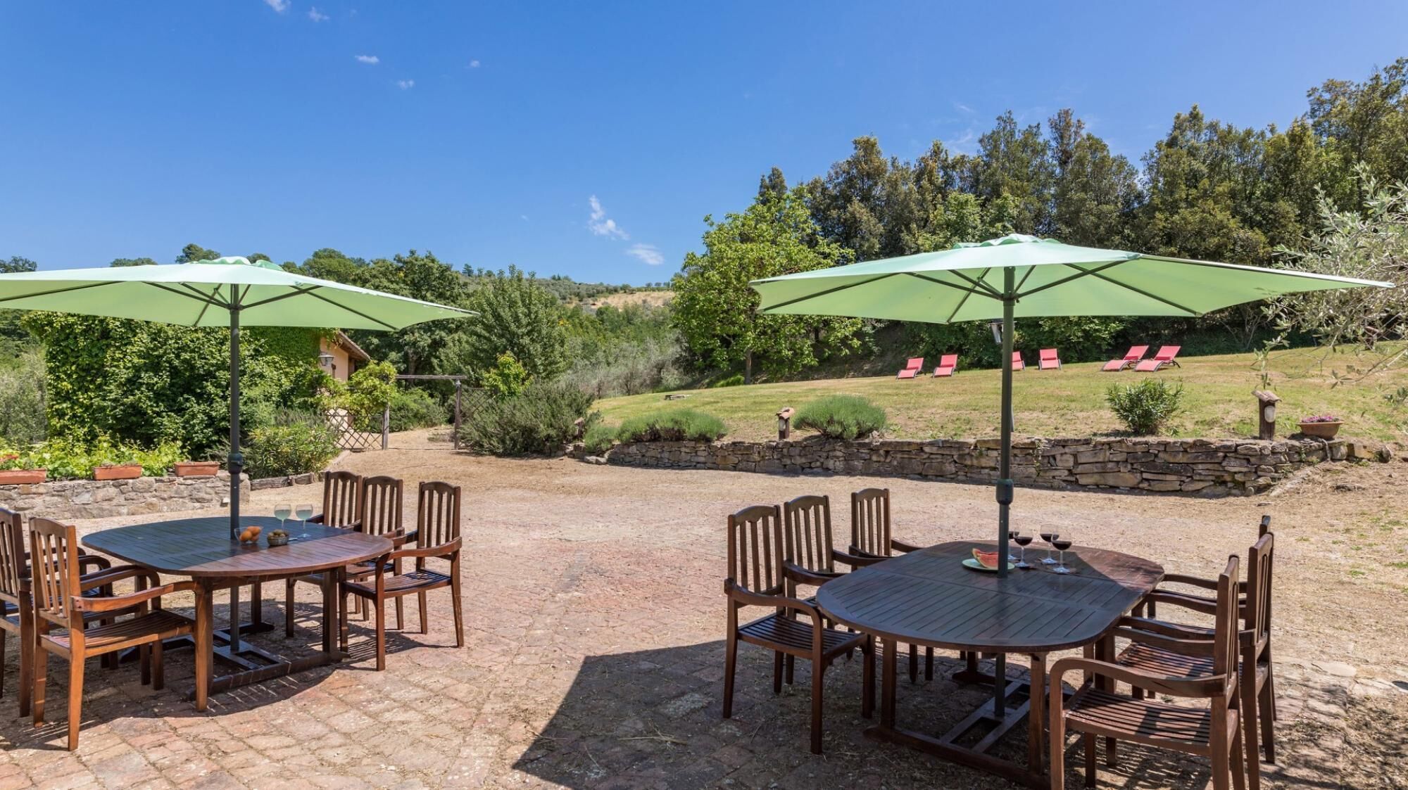 An outdoor patio area with two round tables and chairs, each with a green umbrella. There are stone walls and a grassy hill with trees in the background.