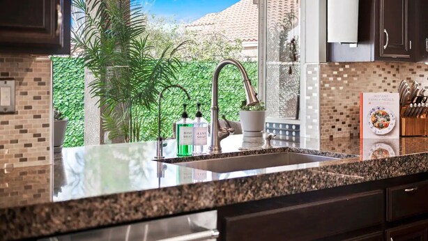 A kitchen with a granite countertop, stainless steel sink, and a window with a view of greenery and a building.