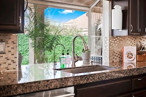 A kitchen with a granite countertop, stainless steel sink, and a window with a view of greenery and a building. - Family friendly home with pool, pool table, games, arcades and equipped kitchen (Indio)