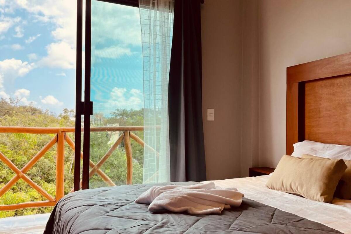 A bedroom with a large bed, a wooden headboard, and a view of greenery through a balcony.