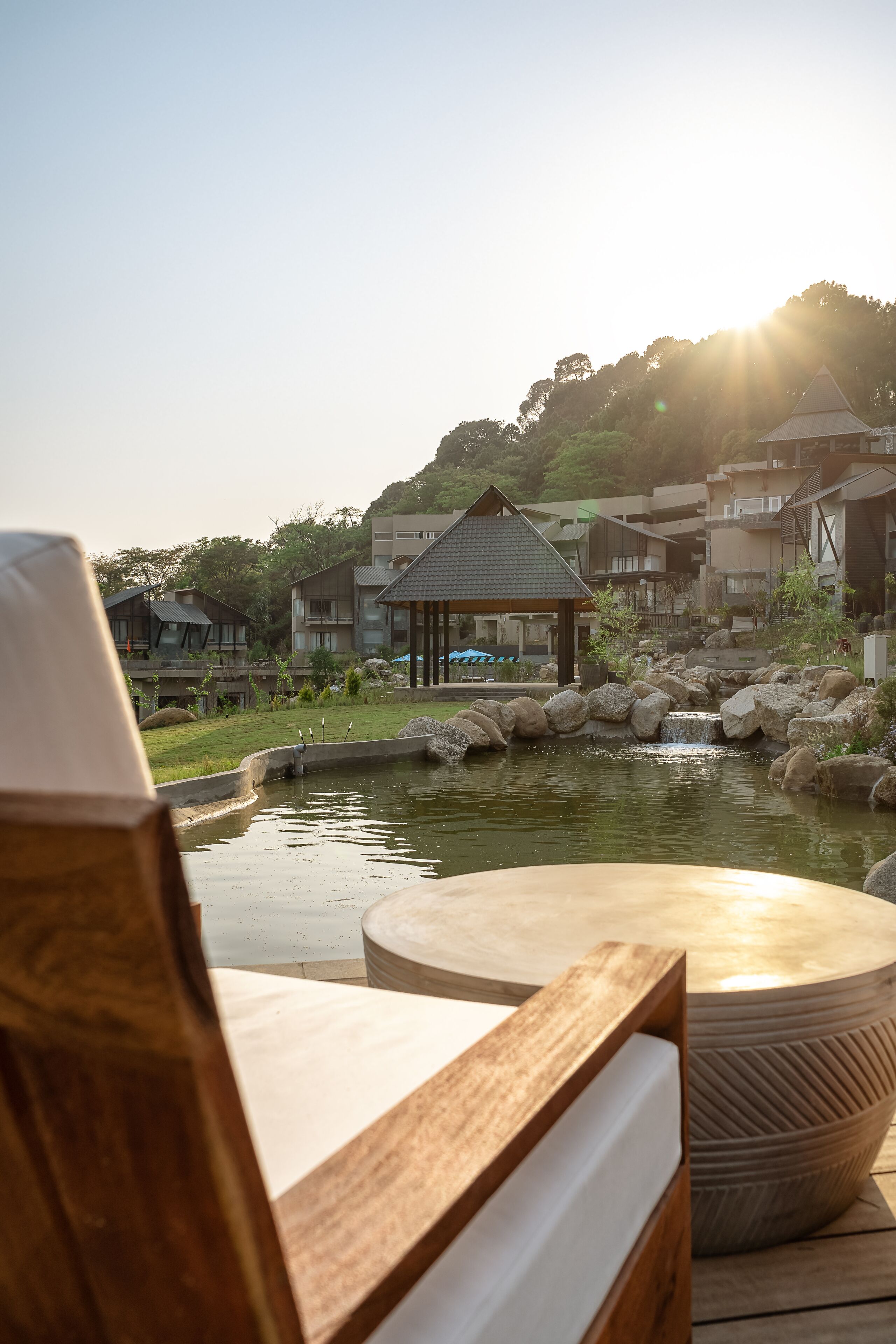 Una terraza de madera con una mesa redonda y una silla, con vista a un estanque con rocas y una pequeña cascada, y edificios al fondo.