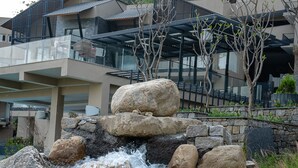 A waterfall cascading over rocks in a landscaped area with a modern building in the background. - Norbu The Montanna, Dharamshala - IHCL SeleQtions (Dharamshala)