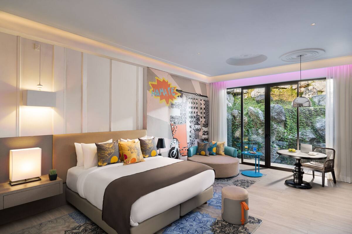 A modern hotel room with a large bed, a desk, a chair, and a view of the outdoors.