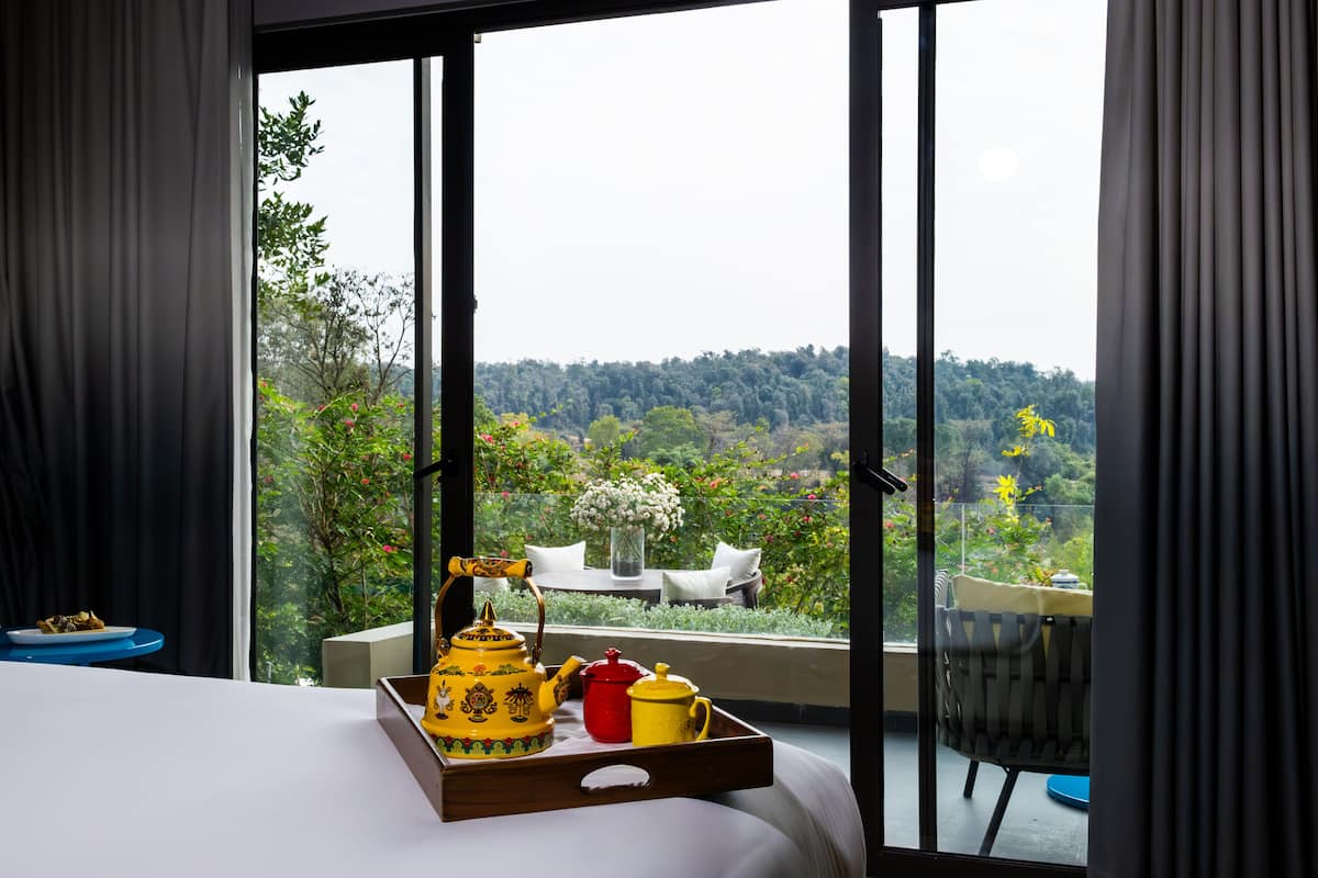 A hotel room with a large window offering a view of a lush garden and distant hills.