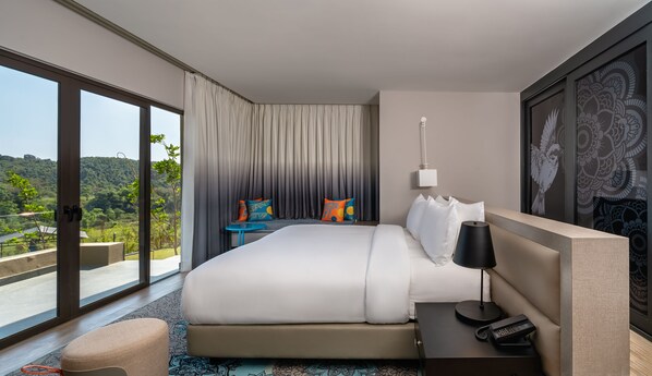 Habitación de hotel moderna con una cama grande, una mesita de noche con lámpara, teléfono y vistas a un paisaje exuberante a través de ventanales de gran altura.