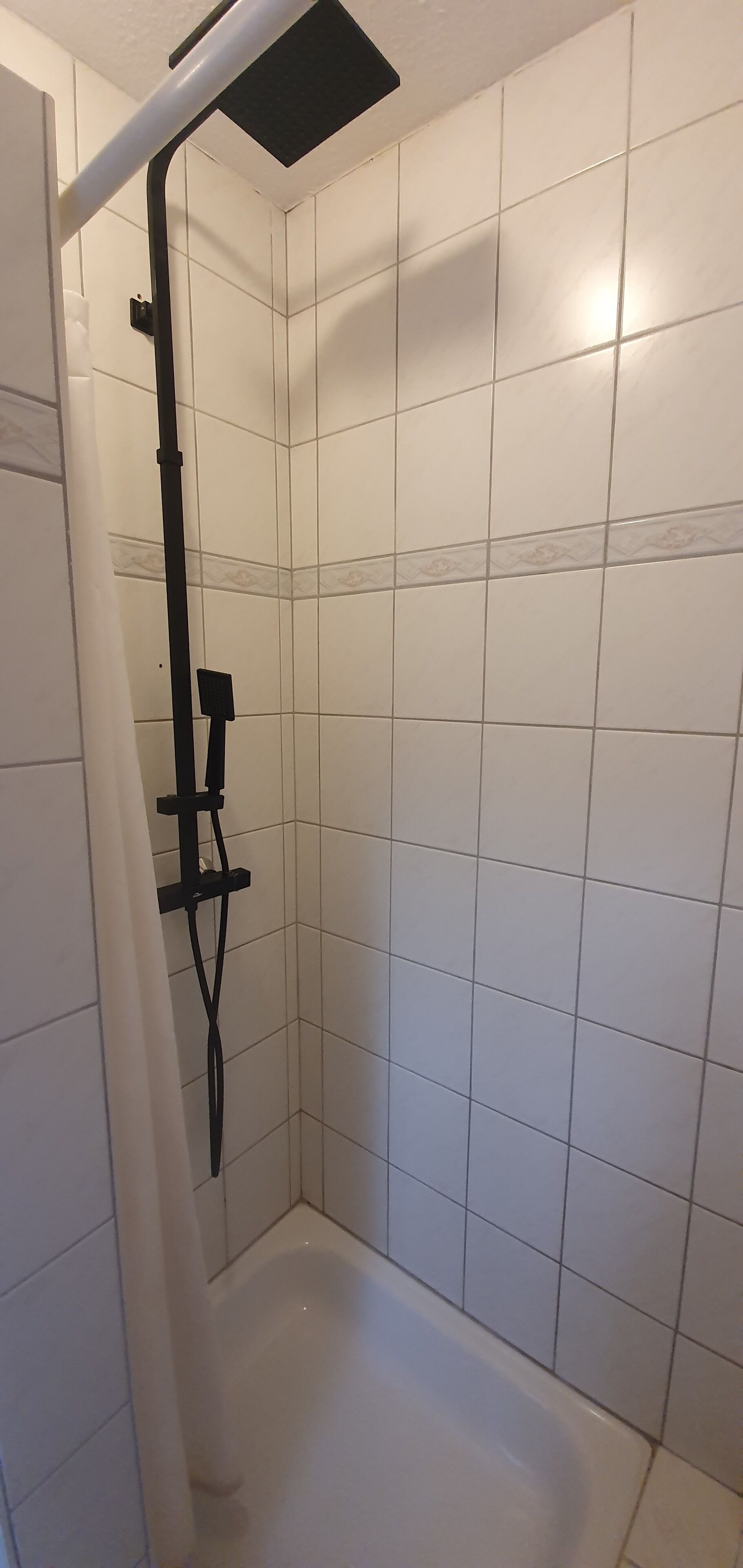 Zona de ducha con paredes de azulejos blancos y un cabezal de ducha negro.