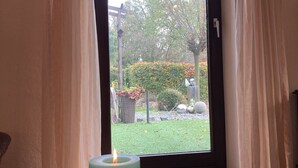 A room with a door leading to a garden. A lit candle is on a wooden tray, and a book is on the floor beside it.