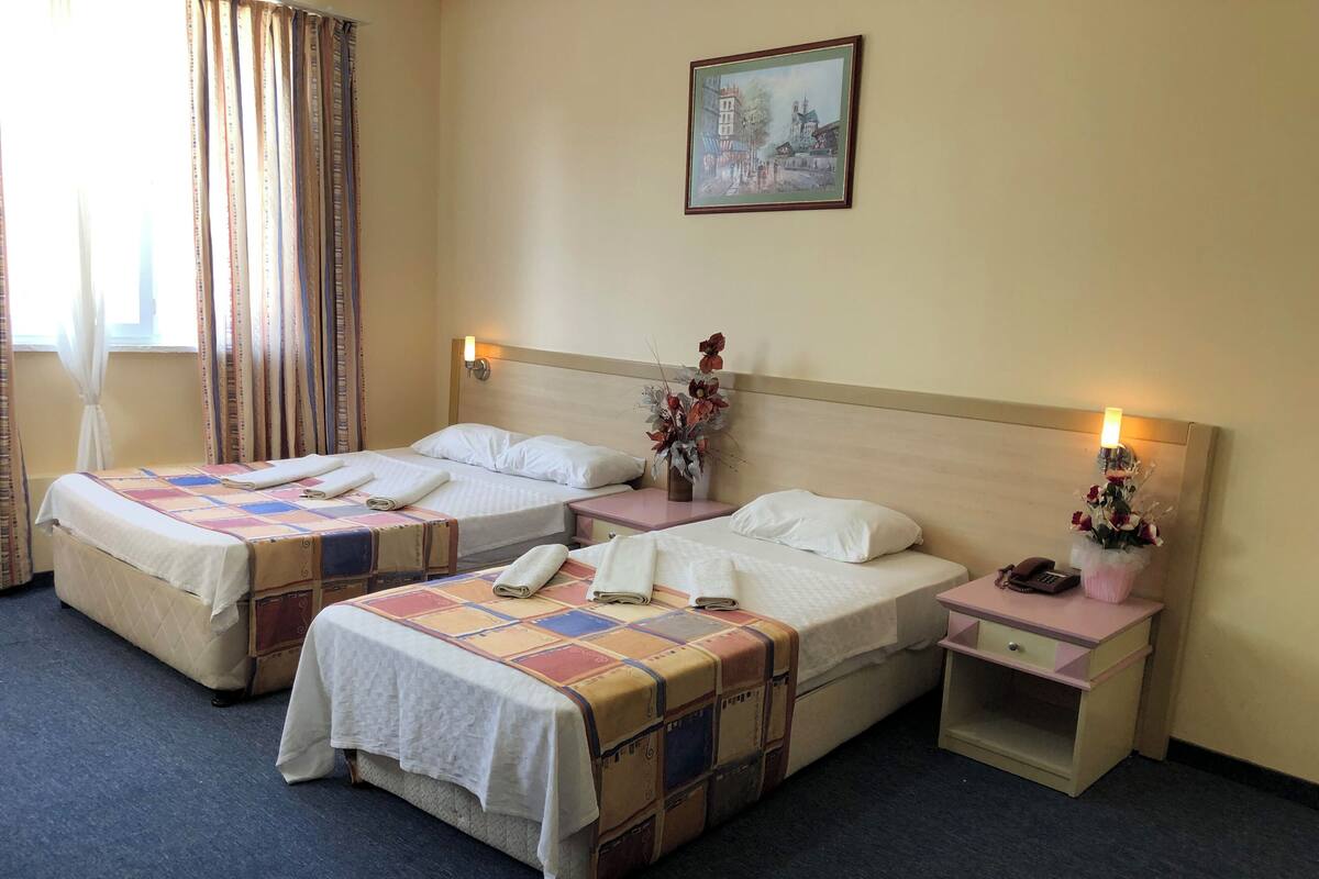 A hotel room with two beds, a bedside table with a lamp, a small cabinet, and a painting on the wall.
