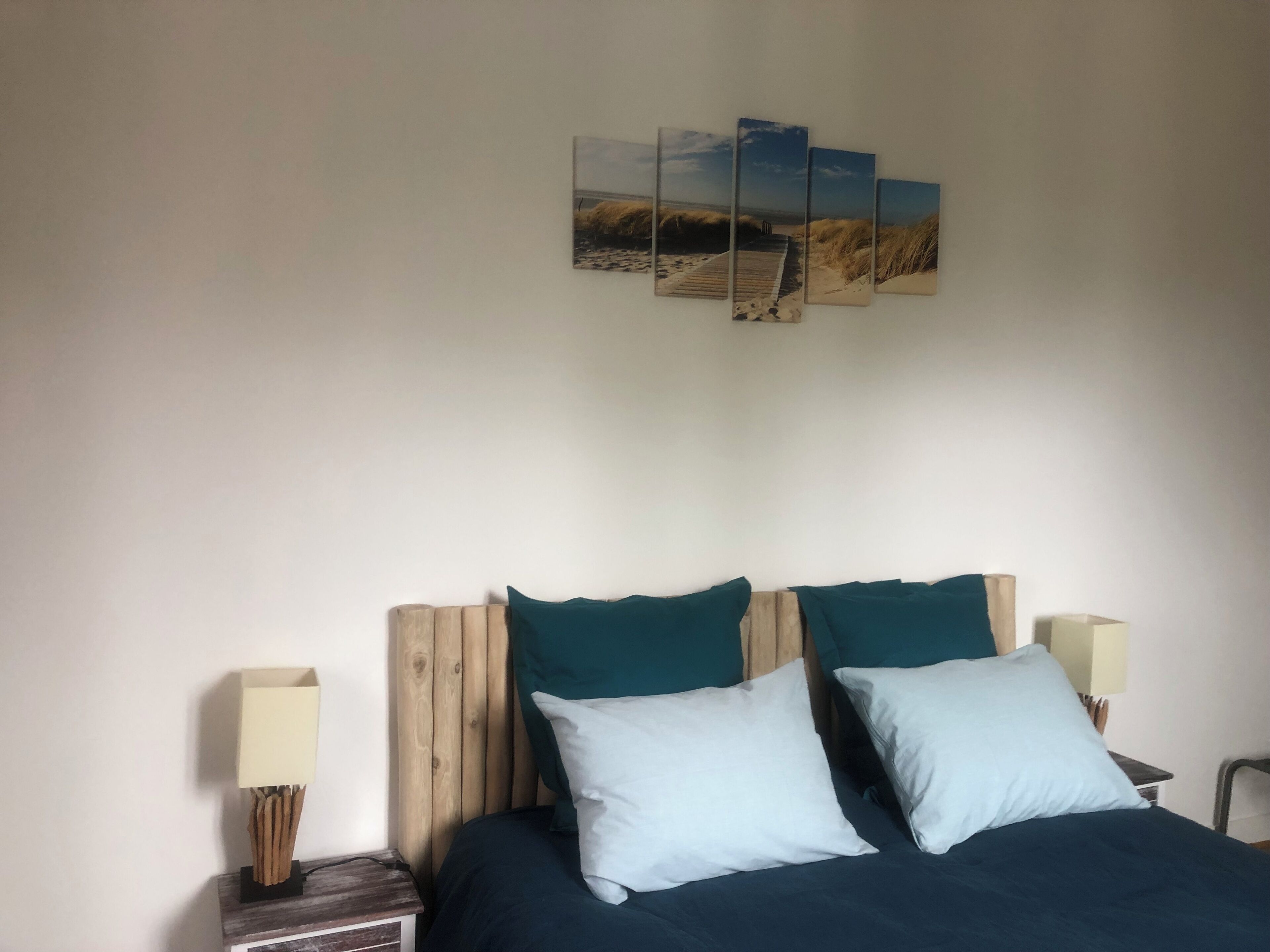 A bedroom with a wooden headboard, two pillows, a bedside table with a lamp, and a wall-mounted artwork of a beach scene.
