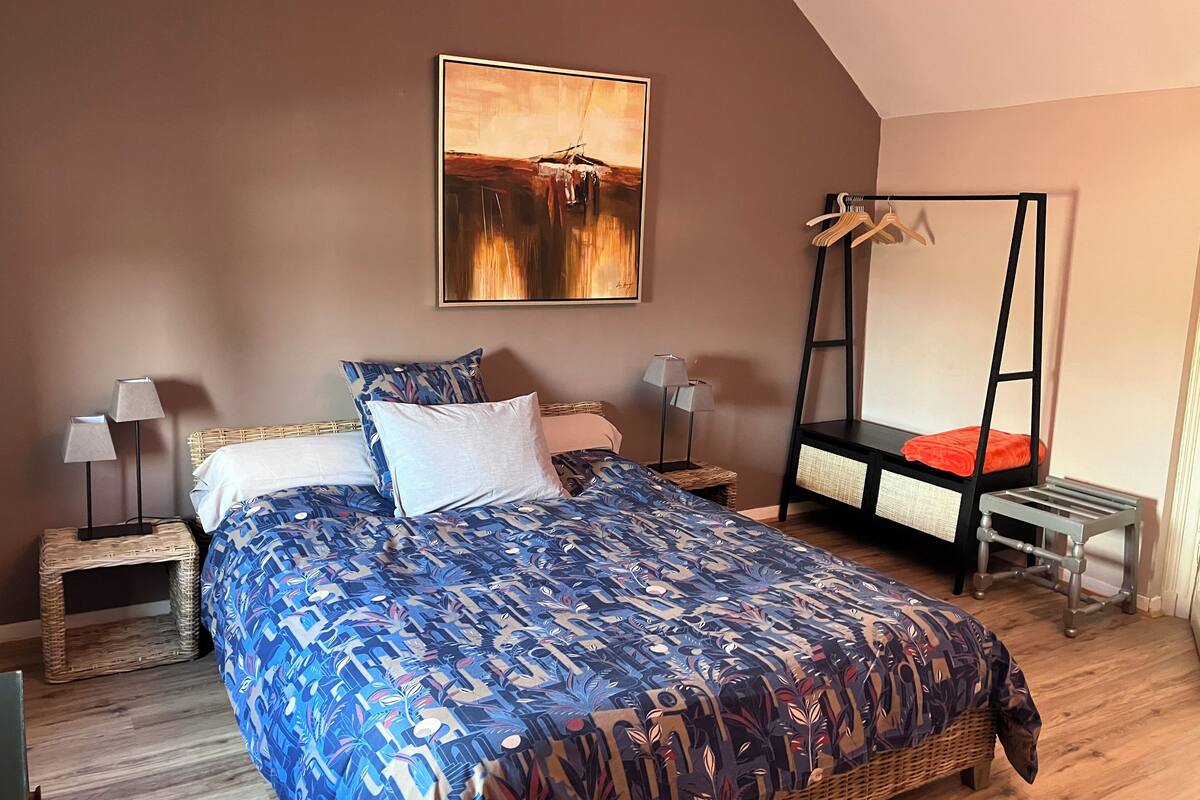 A bedroom with a large bed, two bedside tables, a hanging chair, and a painting on the wall.